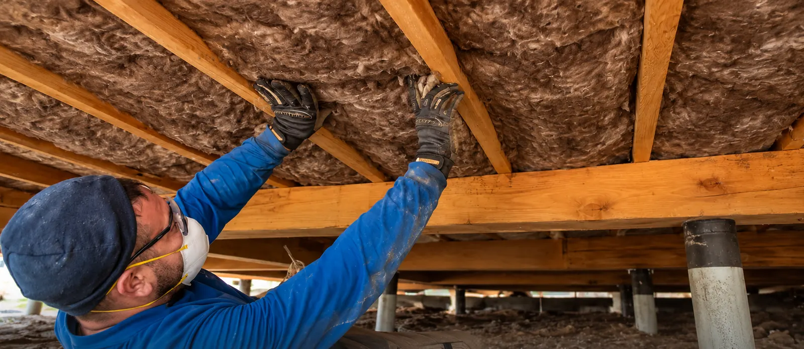 Installer wearing a blue shirt, beanie and respirator fitting brown Earthwool batts between timber underfloor joists